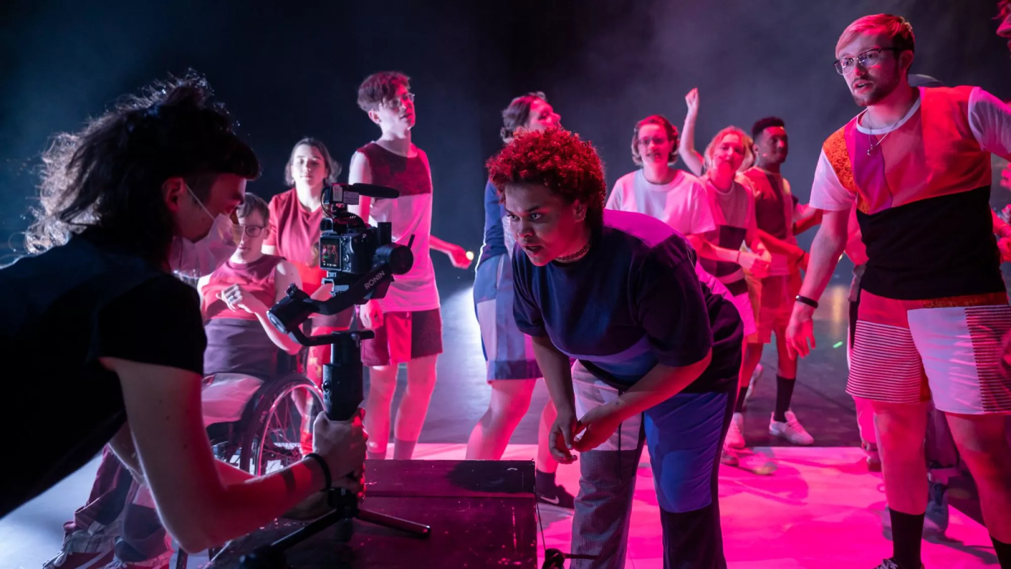 <p>A group of performers stands in pink-blue stage lighting while a camera operator films them up close. Their upcycled costumes combine sporty silhouettes, layered panels, and bold color blocking.</p>