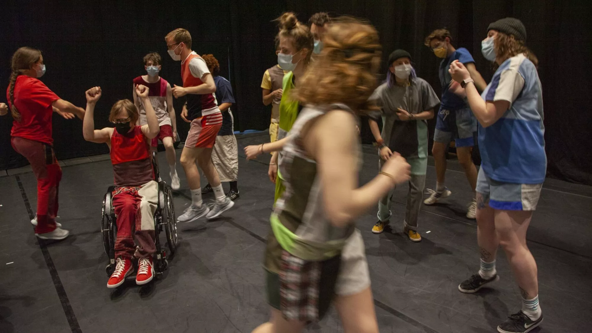 <p>A group of performers moves together across a dark stage, creating a sense of rhythm and collective energy. Their costumes are assembled from upcycled fabric squares, forming sporty silhouettes with vivid color blocking.</p>