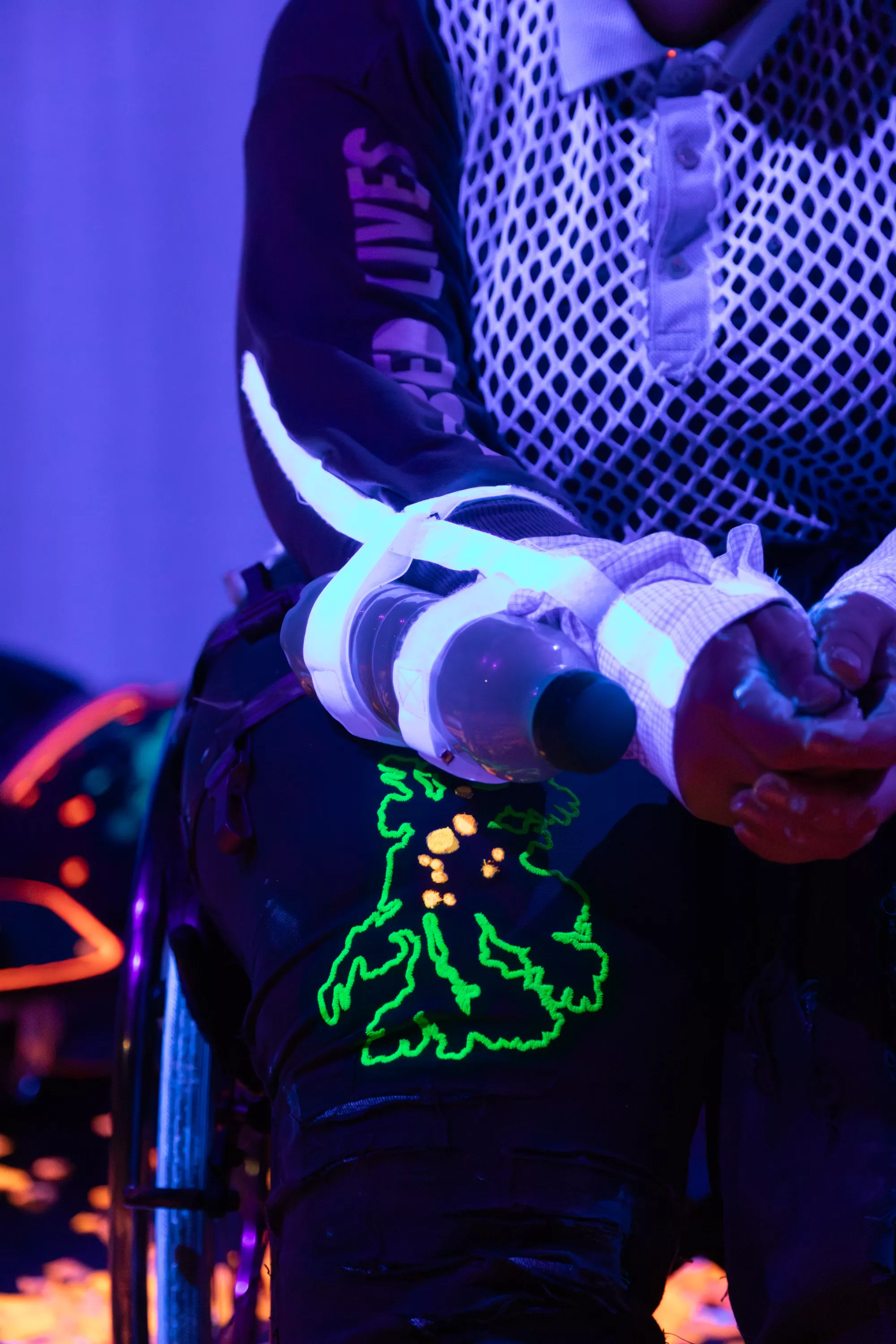 A close up of a performer using a wheelchair. Under blue and purple stage light, neon green and orange details on their clothing appear to glow, echoing the bioluminescent atmosphere of the performance. There is a bottle attached with withe straps to the forearm of the performer filled with seeds that make a sound when they move the arm. They are wearing a top from white net fabric that is glowing in the UV-light.