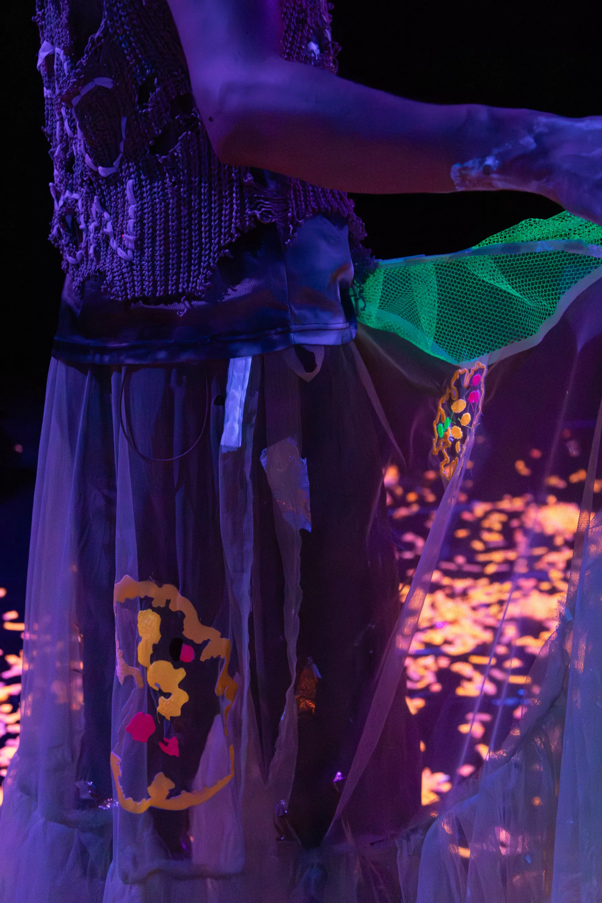 Close-up scene of a performer. A performer’s torso and outstretched arm are lit in purple and blue stage light. They wear a knitted torn top with a text stitched on it and a transparent layered skirt with glowing embroidered lichen in yellow, pink, and orange. A green mesh fabric extends from their arm, while blurred orange confetti is glowing across the dark stage in the background.
