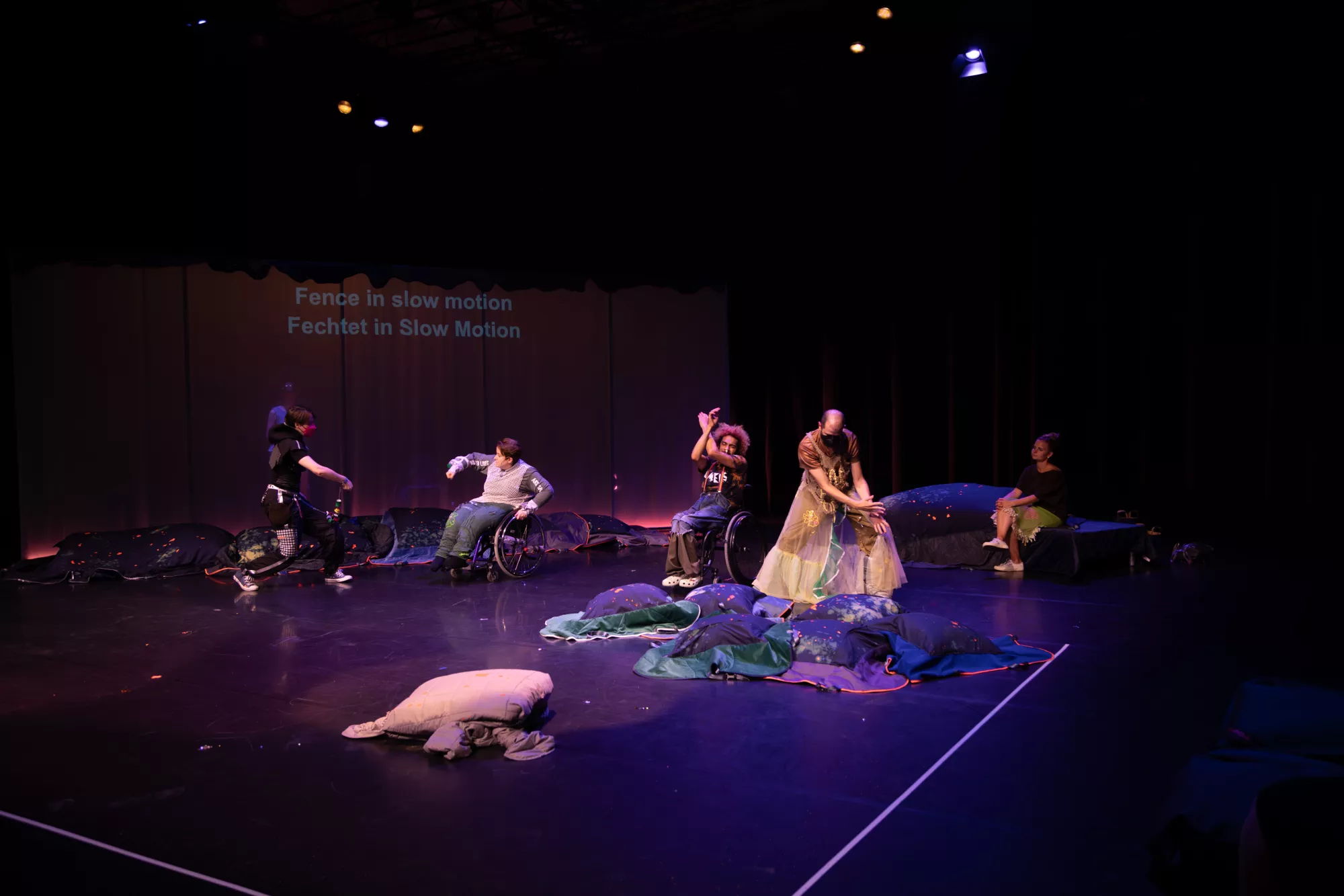 Four performers are dancing across a dark stage lit with purple and blue light. Some performers use wheelchairs, while others stand or sit among soft, pillow-like textile sculptures. The performers are fencing in slow motion, echoing the projected text on the backdrop: “Fence in slow motion / Fechtet in Slow Motion.” The stage is arranged with dark cushions, fabric objects, and scattered small materials, creating a dreamlike performance environment.