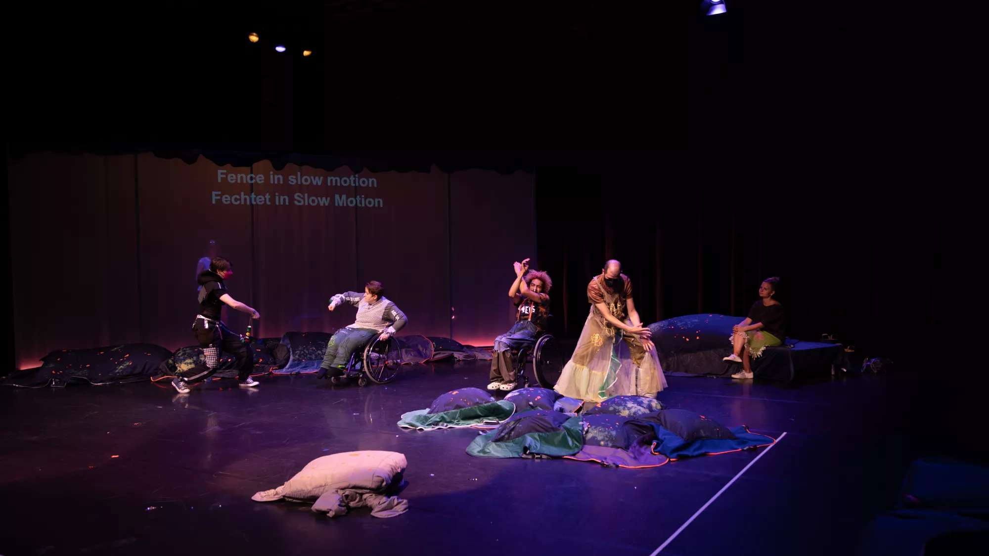Four performers are dancing across a dark stage lit with purple and blue light. Some performers use wheelchairs, while others stand or sit among soft, pillow-like textile sculptures. The performers are fencing in slow motion, echoing the projected text on the backdrop: “Fence in slow motion / Fechtet in Slow Motion.” The stage is arranged with dark cushions, fabric objects, and scattered small materials, creating a dreamlike performance environment.