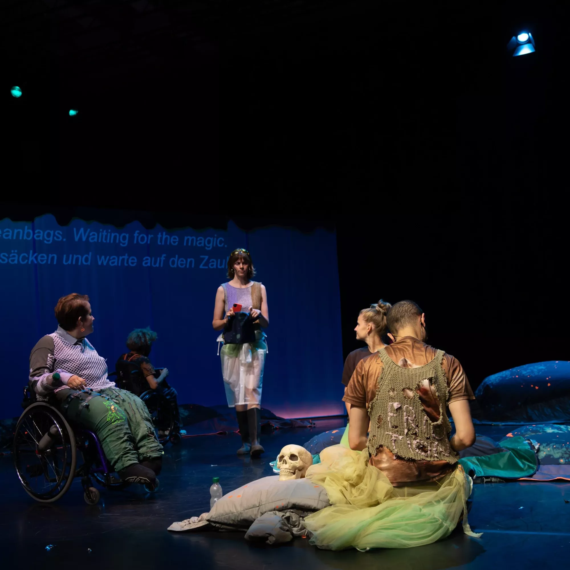 Five performers are on a dark stage lit with blue and green light. Two performers are using a wheelchair, two performers sit or kneel on the floor, and another stands in the centre holding an object. The stage is scattered with cushions, fabric, a skull prop, and soft sculptural elements, while projected text appears faintly on a curtain in the background.
