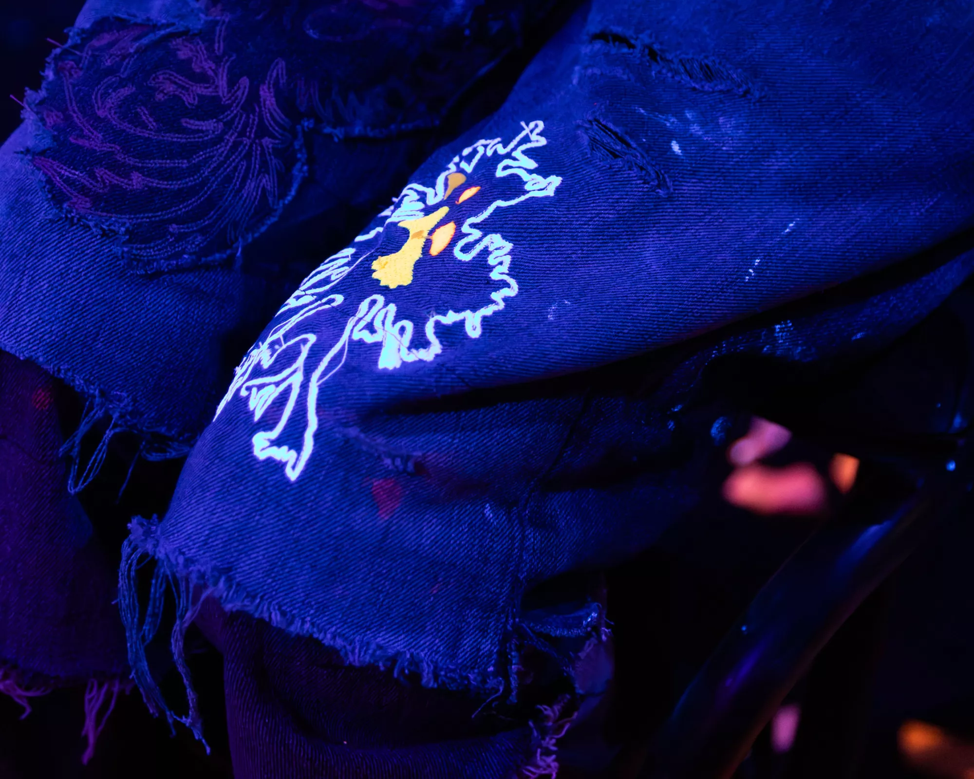 Close-up scene of a performer. Blue and purple stage light illuminates a performer’s layered, torn denim trousers. A glowing, embroidered phoenix motif appears on the fabric, with white outlines and small yellow-orange details. The surrounding stage is dark, with blurred orange and pink light effects in the background.