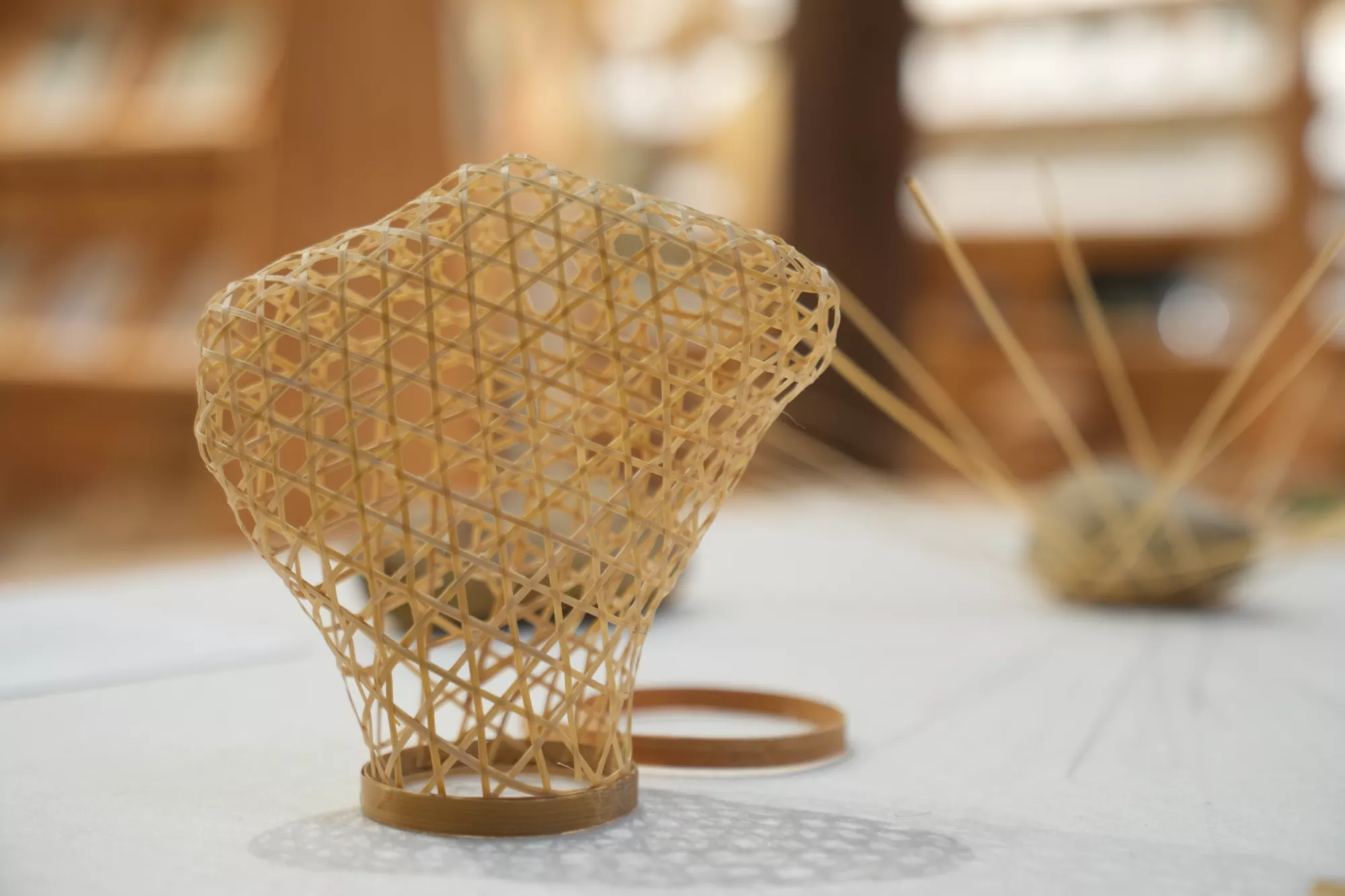 <p>A bamboo brooch displayed on a white surface. The brooch is woven from thin bamboo strips into an open, irregular, sculptural shape. It was made by weaving the bamboo around a stone so that the finished piece takes on the stone’s contour. Another bamboo element and fine strips appear blurred in the background.</p>