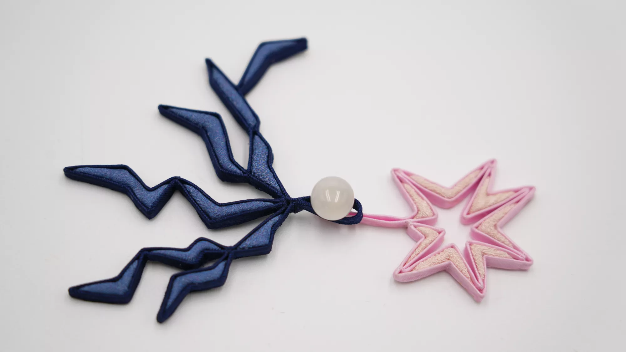 <p>A finished pankou design on a plain white surface. A dark blue knot-like form with angular, branching loops is connected to a pink star-shaped loop by a round white bead. The piece combines structured corded edges with translucent and cream-coloured textile inserts.</p>