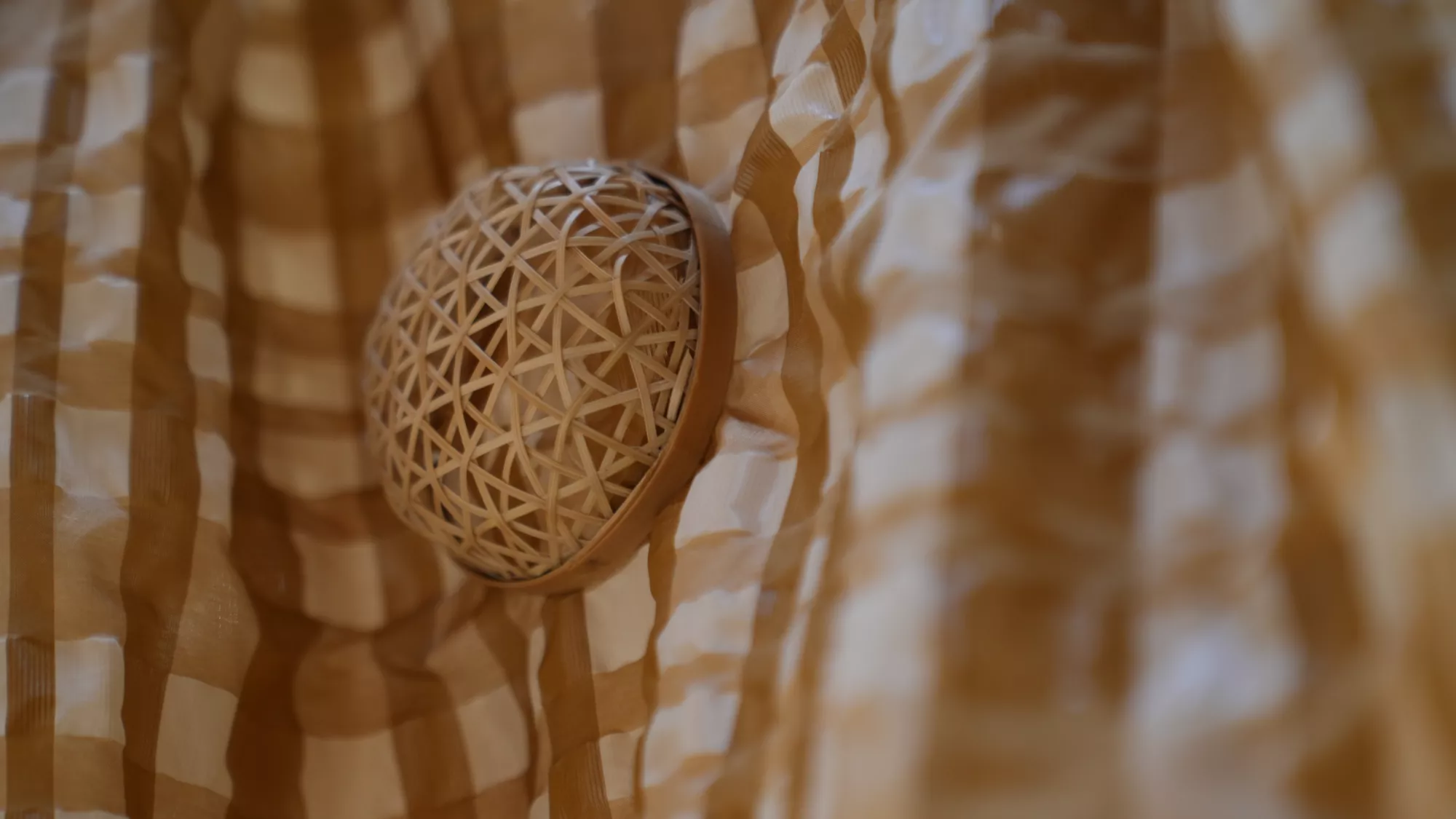 Close-up of a bamboo brooch attached to a brown and cream checkered skirt. The brooch has a rounded, dome-like shape made from interlaced strips of bamboo, creating an open woven pattern. The warm tones of the bamboo complement the structured checkered fabric of the skirt.