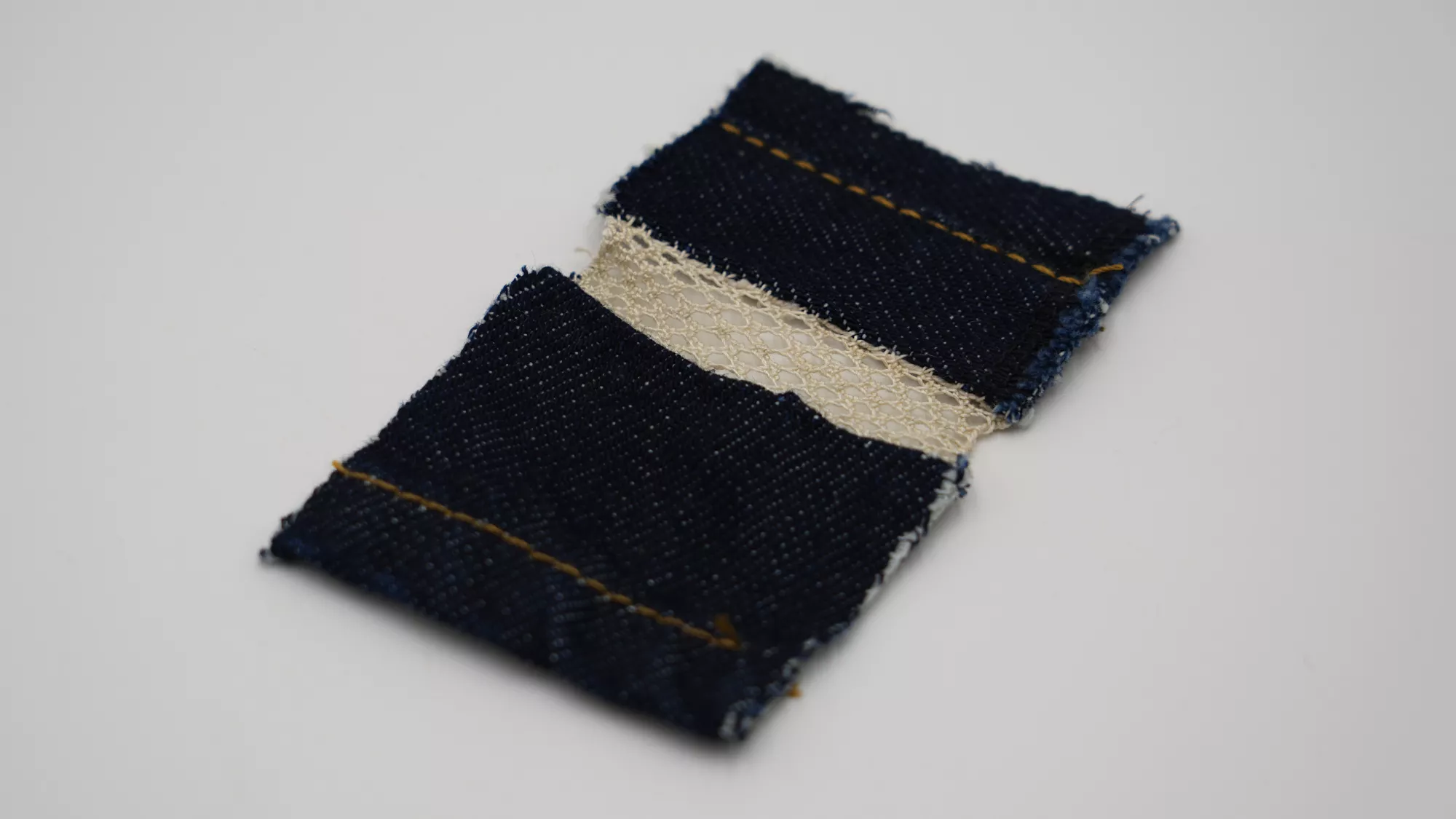 Close-up of a Xiaoshan lace textile sample on a plain white surface. Two dark blue jeans scraps are connected by a narrow strip of cream-coloured lace. The denim edges are slightly frayed, and visible brown stitching runs across the fabric, emphasizing the handmade construction.