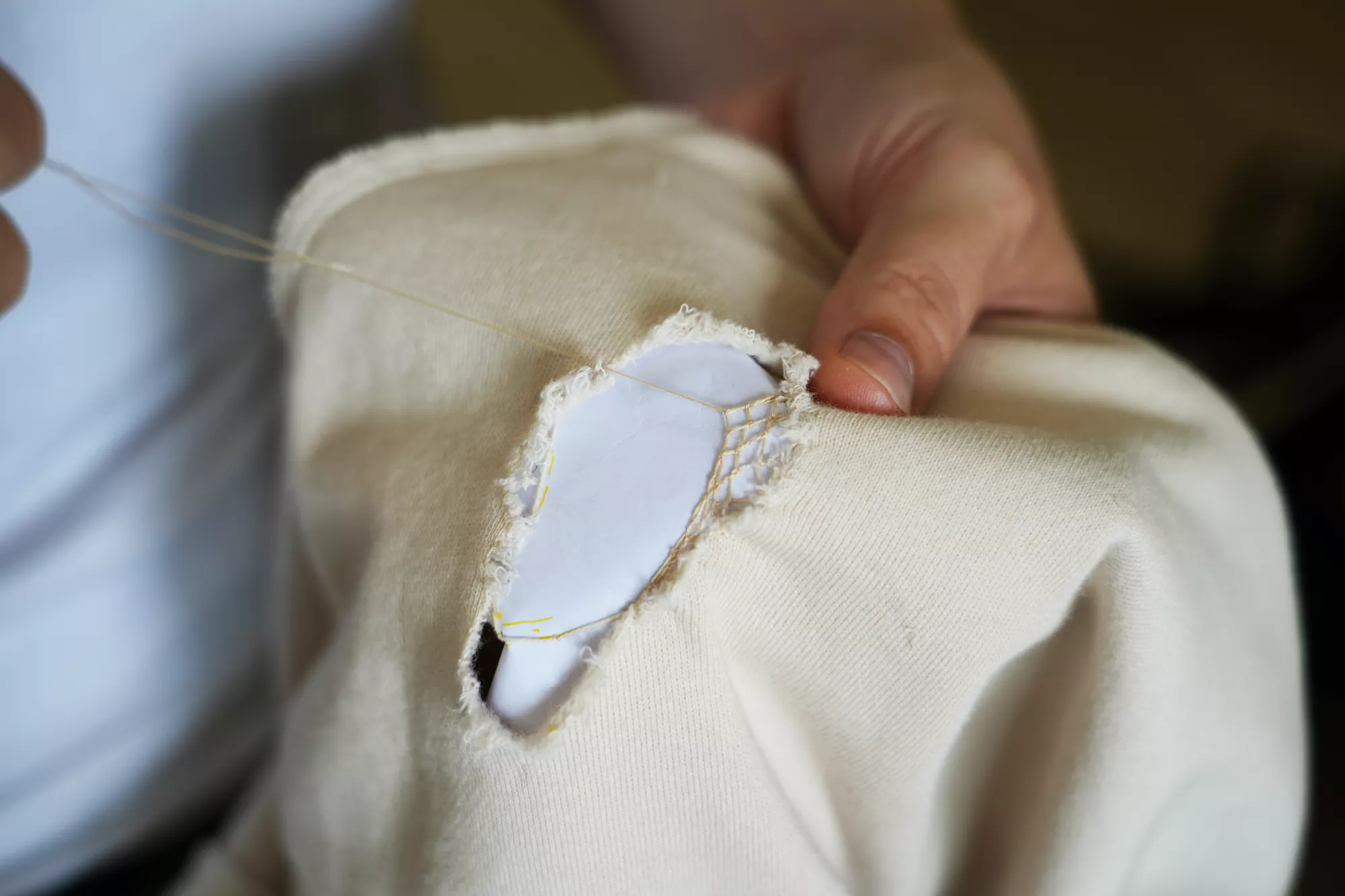 Close-up of a broken cream-coloured sweater with a hole in it, repaired using Xiaoshan lace. A hand stitches fine beige thread across the frayed opening, creating a small lace-like mend over a white backing material.