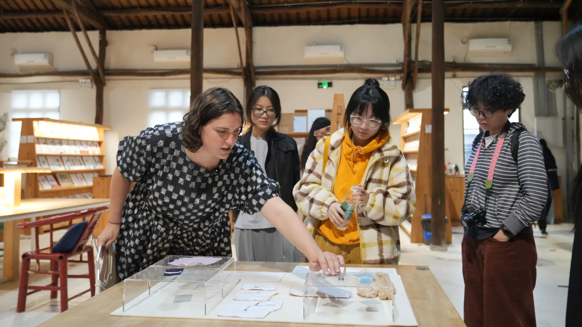 Nicole Frei presents textile samples to a group of visitors at Rong Design Library. She leans over a table and points to small material experiments displayed under clear acrylic boxes, including pieces of Xiaoshan lace and fabric samples. The visitors stand closely around the table, observing the objects in a spacious room with wooden beams, shelves, and warm lighting.