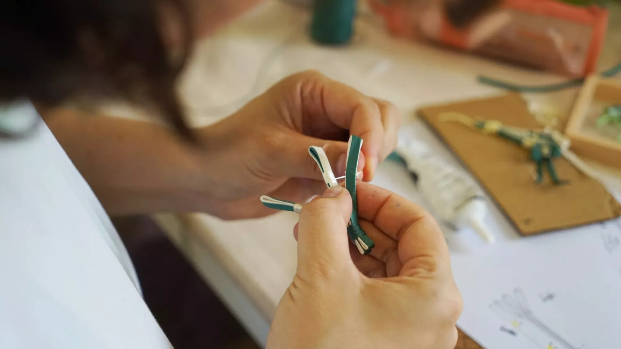 A close-up showing a person working on sewing together a pankou. The Pankou is made of dark green and cream strings.
