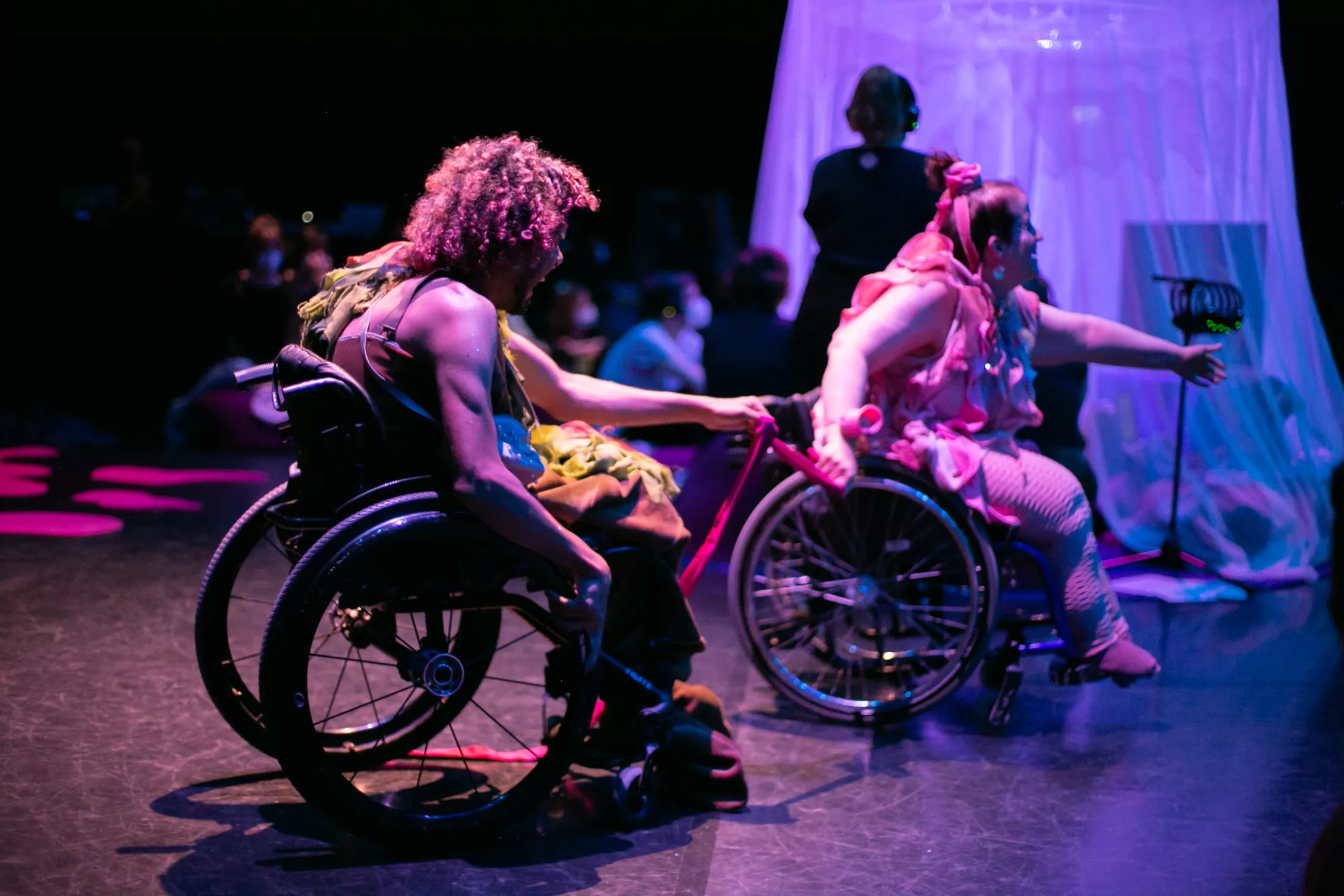 A white and a black performer using wheelchairs move across a dark stage under pink, purple, and blue lighting. They are connected by a pink strap or fabric band, with one performer reaching towards the other while the second extends an arm outward. A translucent fabric tent that looks like a jellyfish and blurred audience members are visible in the background, creating an immersive performance atmosphere.