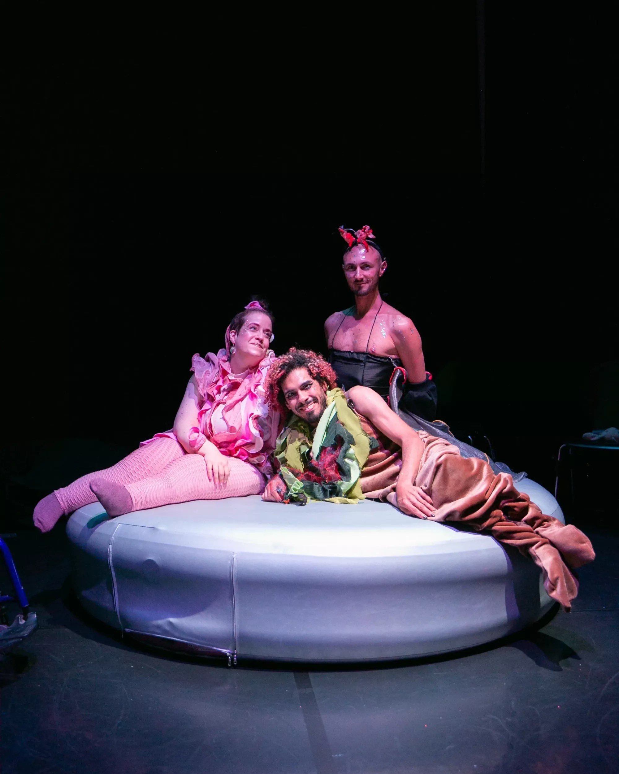 Scene from Pleasure Creature Comforts by Criptonite. Three performers pose together on a large, round, pale inflatable platform in a dark stage space. They wear colourful, layered costumes in pink, green, black, and brown, with sculptural accessories and theatrical makeup. The group leans closely together under soft stage lighting, creating an intimate and playful tableau.