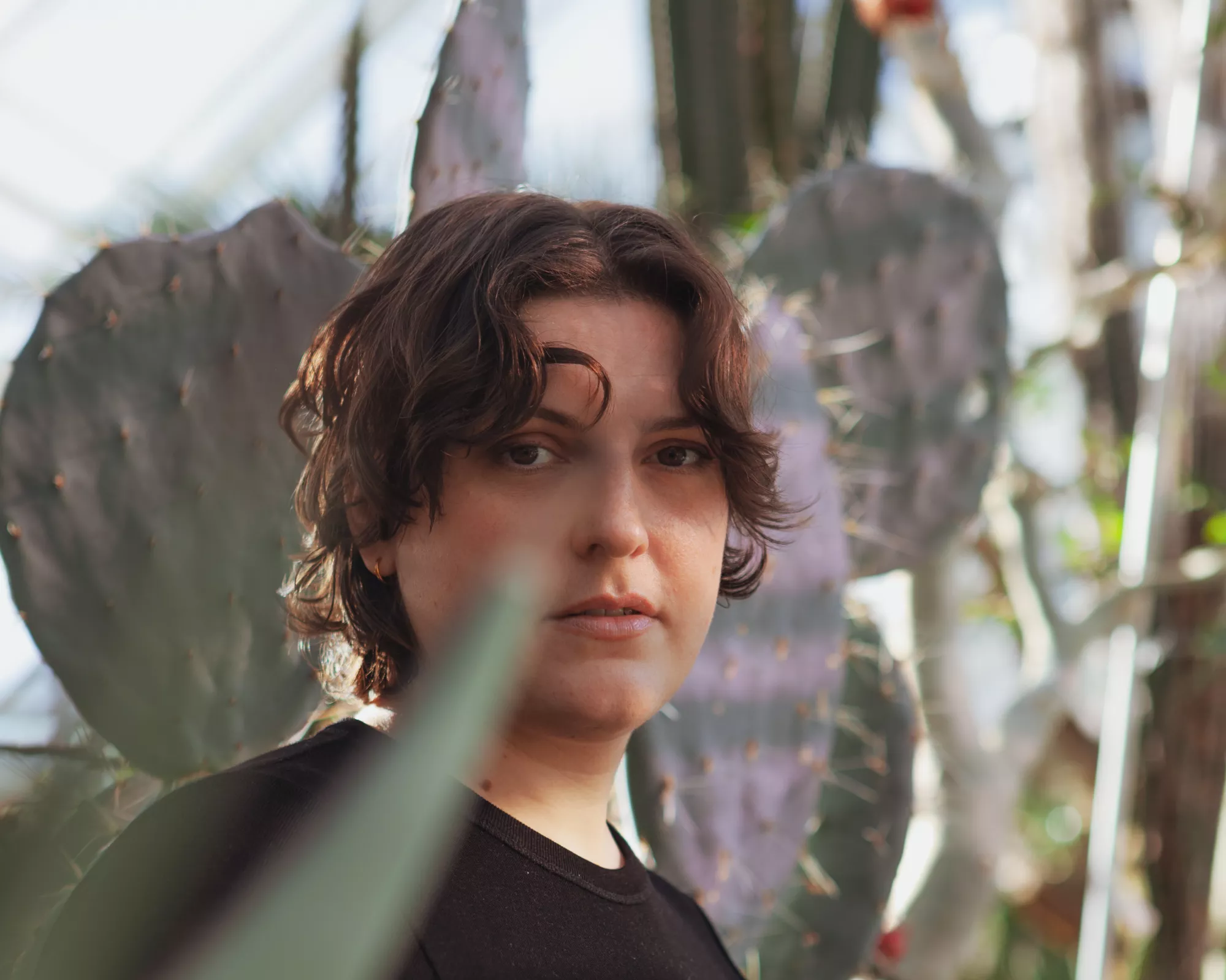 <p>Portrait of Nicole Frei. She is a white female, with wavy brown, shortish hair. She is standing among large cacti and succulent plants, wearing a black shirt and looking directly at the camera. The foreground and background are softly blurred by long plant leaves and cactus forms.</p>