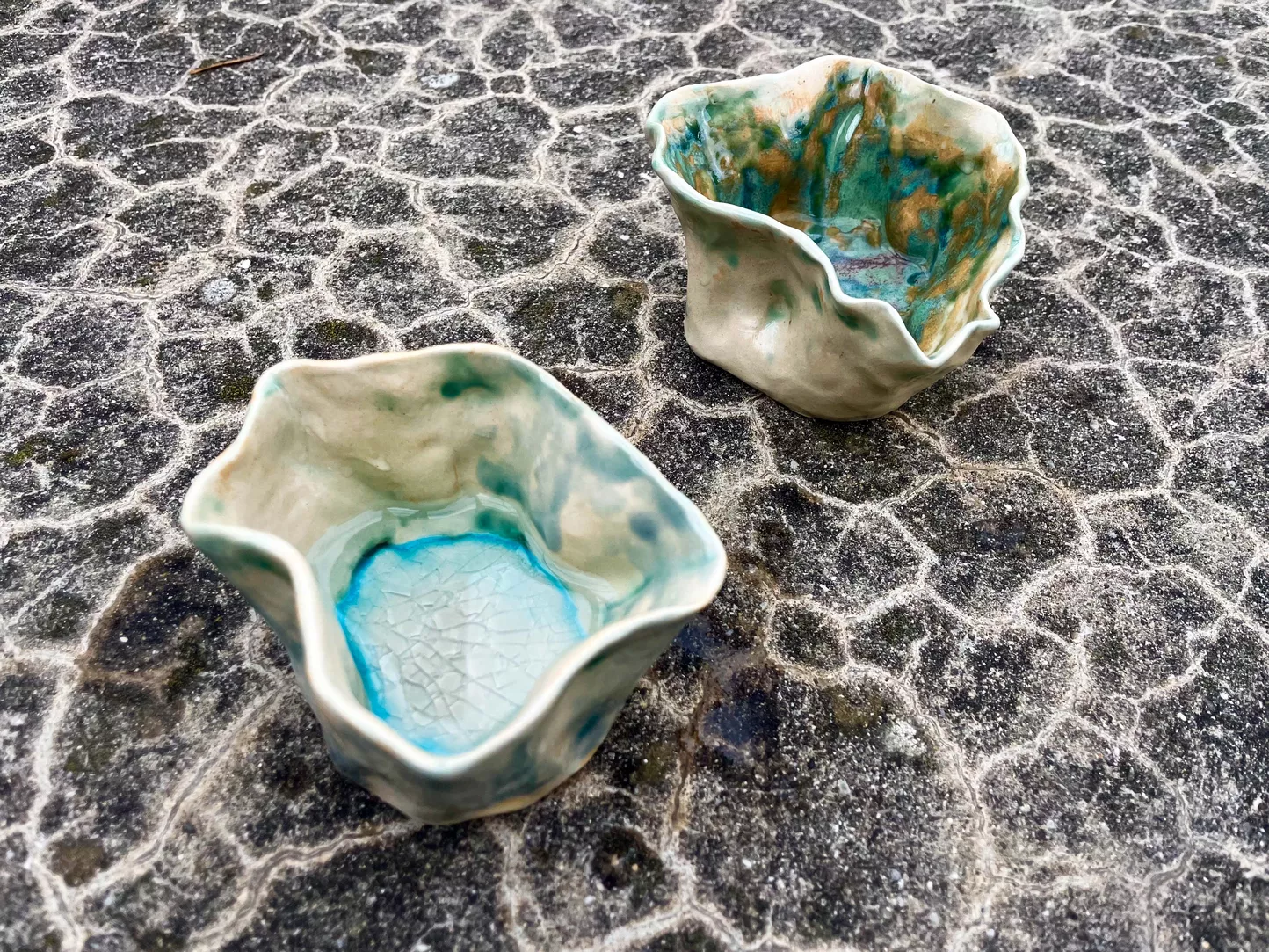 Two small handmade ceramic vessels by Anna-Tia Buss from From Texture to Form Exploration sit on a dark, patterned stone surface. The vessels have irregular, softly folded edges and glossy glazes in pale cream, turquoise, green, and earthy brown tones. The closer vessel has a light blue pool of glaze at the bottom with a fine crackled texture, while the second vessel shows more saturated green and brown glaze marks inside.