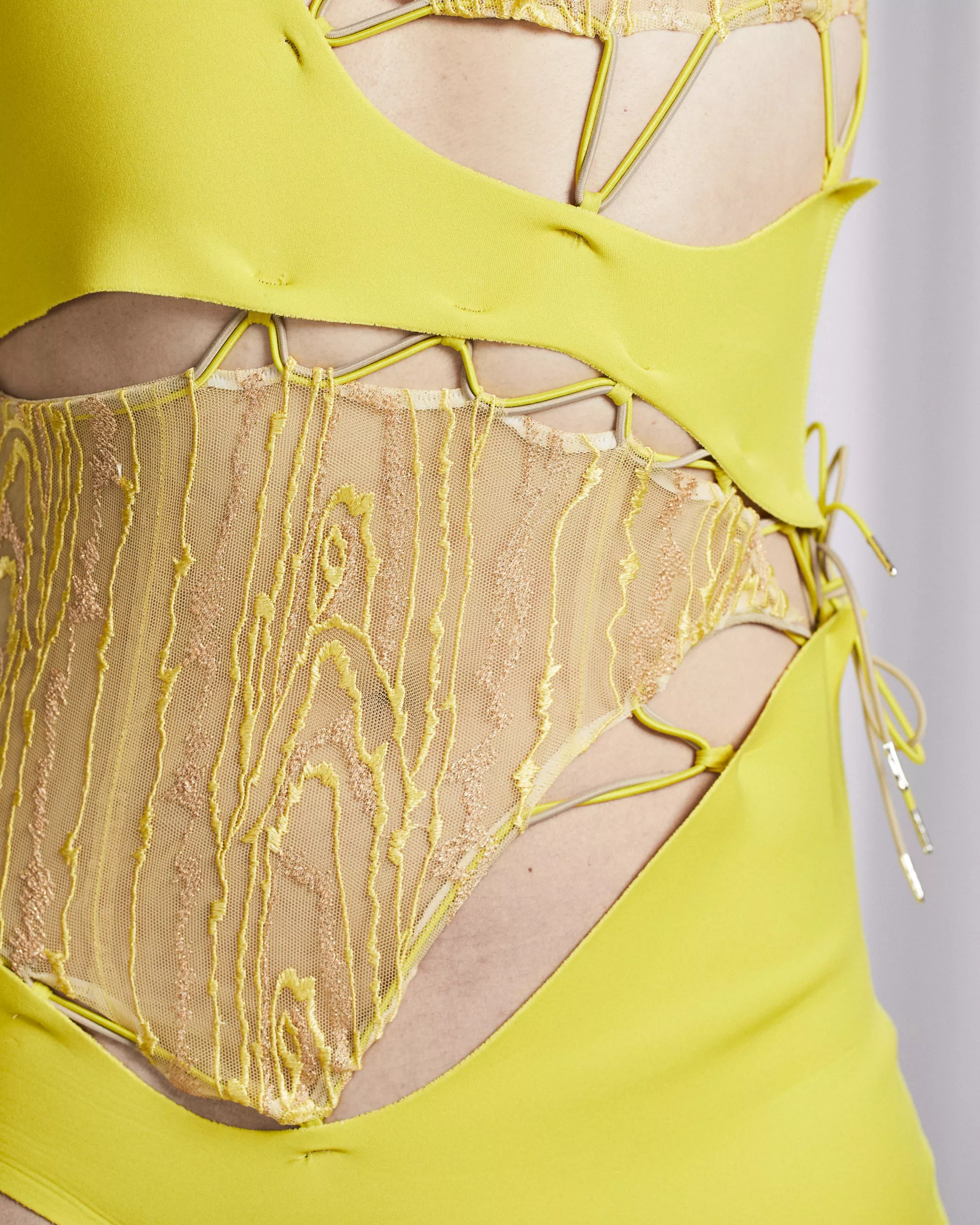 <p>A Close-up of a white skinned person wearing a bright yellow neopren garment with cut-out sections connected by corded loops and lacing. Transparent yellow-orange lace panels with embroidered, organic wood patterns are inserted between the openings, creating a layered textile construction.</p>
