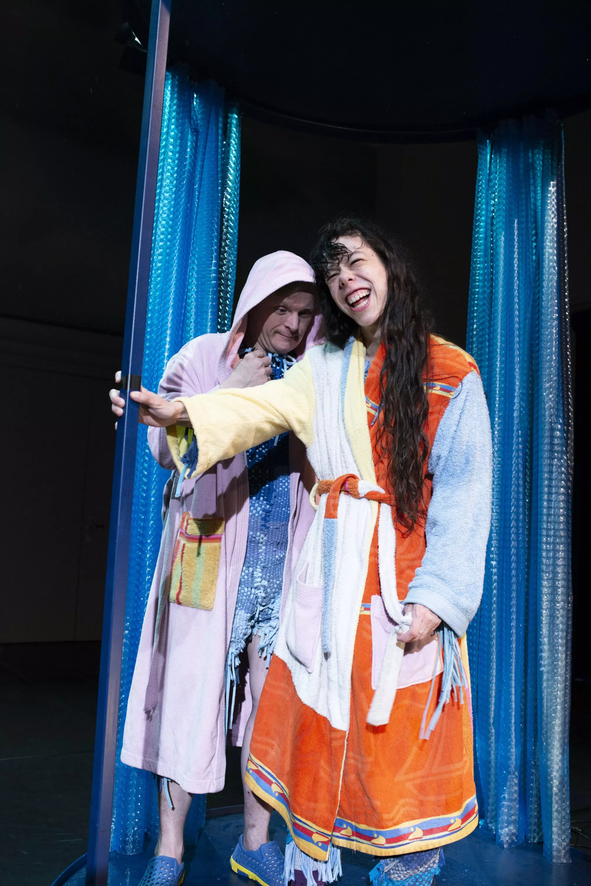 Two performers stand between shimmering blue curtain panels in a stage setting. They wear colourful bathrobes made from old towels and patchworked together in pale pink, light blue, orange, and blue. One performer reaches toward a vertical pole while smiling widely; the other stands slightly behind them.