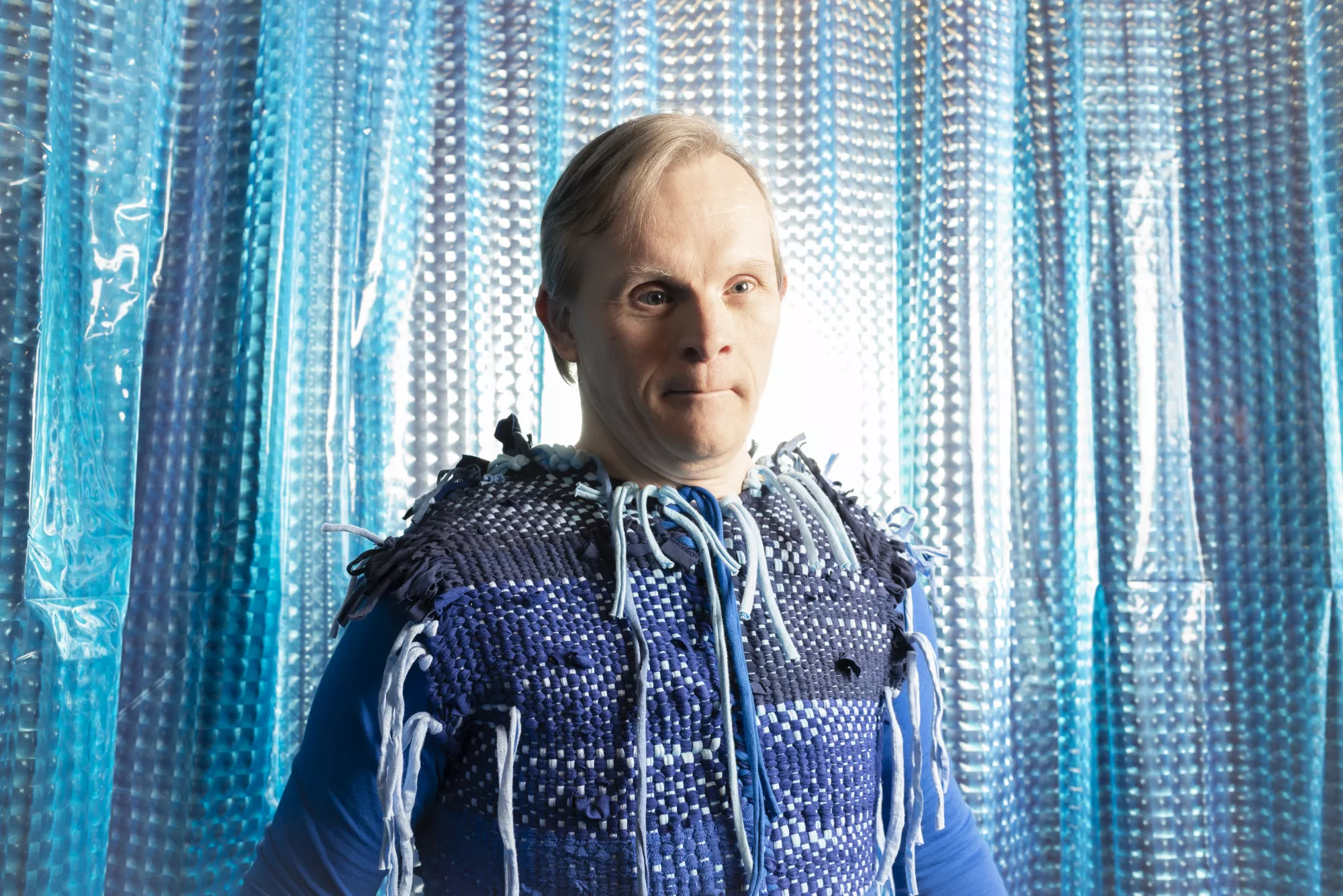 A portrait of a white-skinned performer, who stands in front of a shimmering blue and silver curtain, wearing a blue woven textile costume with light blue cords and loose strands hanging from the shoulders and chest. Bright backlighting creates a glowing effect around the performer and reflects across the textured curtain, emphasizing the layered blue tones of the costume and stage environment.