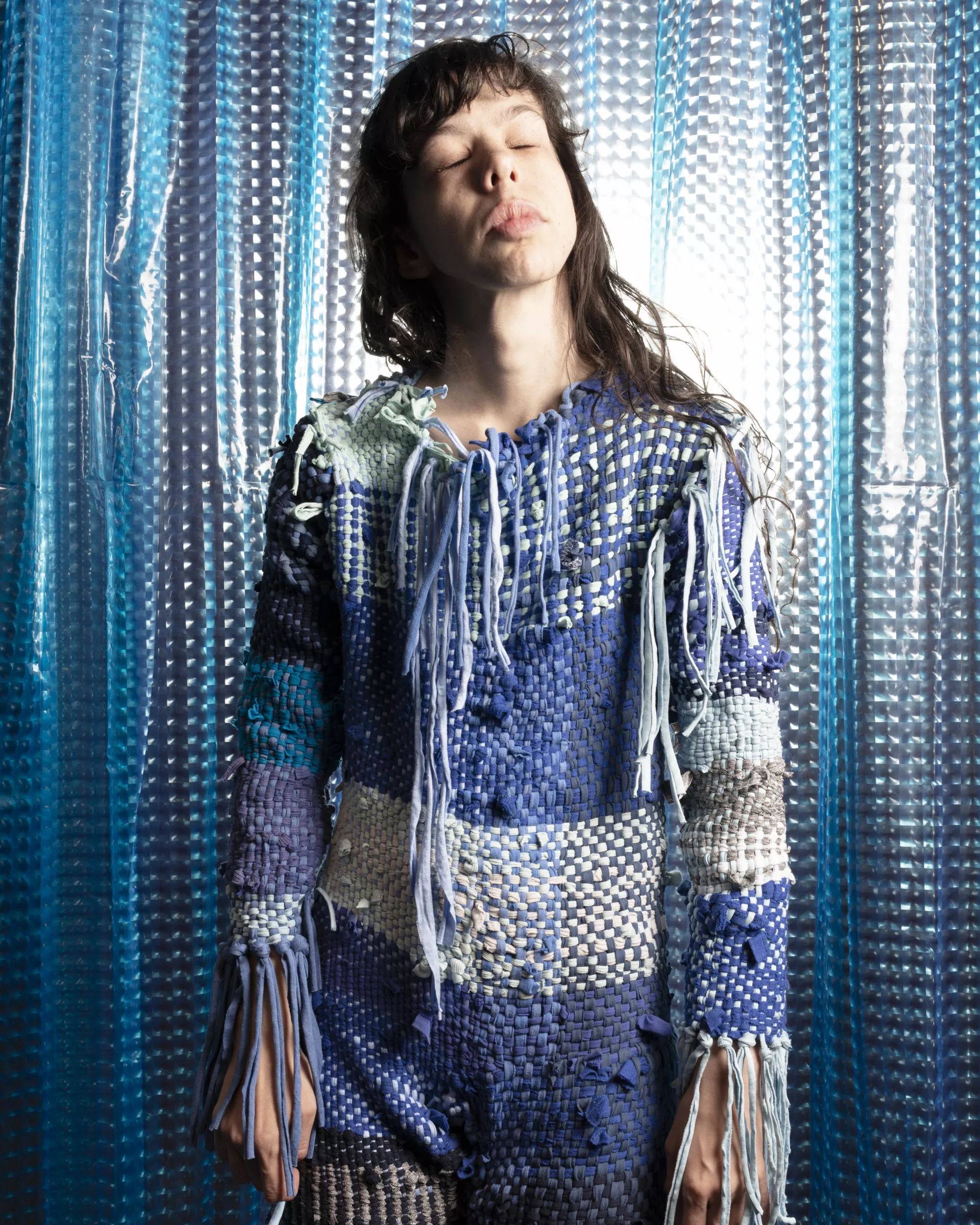 A white-skinned performer stands in front of a shimmering blue and silver curtain, wearing a full-body woven textile costume in shades of blue, grey, cream, and pale green. Long loose strands hang from the sleeves and chest, adding movement and texture to the garment. The performer’s eyes are closed and their face is lifted slightly toward the light, creating a quiet, reflective mood within the staged setting.