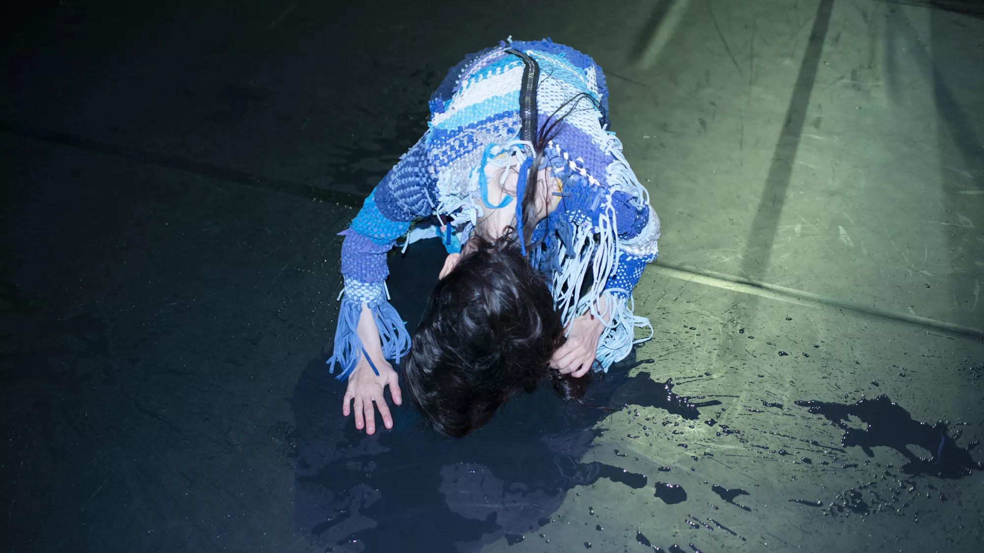 <p>A performer with dark brown hair in a blue woven costume is curled forward on a black dance floor, surrounded by reflective puddles of water. Long loose textile strands hang from the costume.</p>