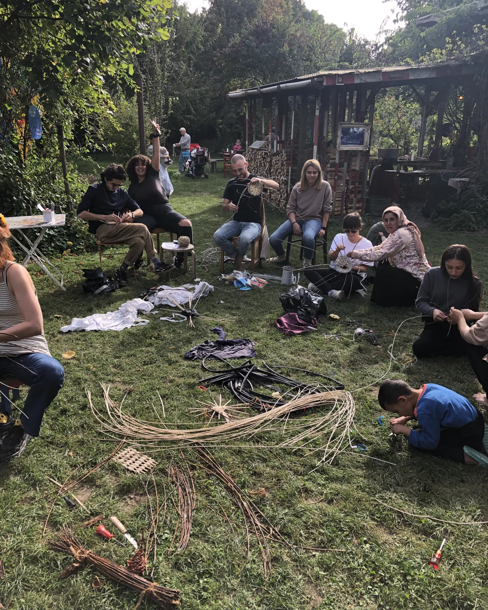 Basket weaving workshop by Katharina Berger at Hardturm Brache in Zurich. A group of people sit and work together on the grass, using branches, reeds, cord, fabric, and other found materials to make woven and constructed objects. Materials are spread across the ground, while participants work individually and in small groups in a green garden-like setting with trees, tables, and a wooden shelter in the background.