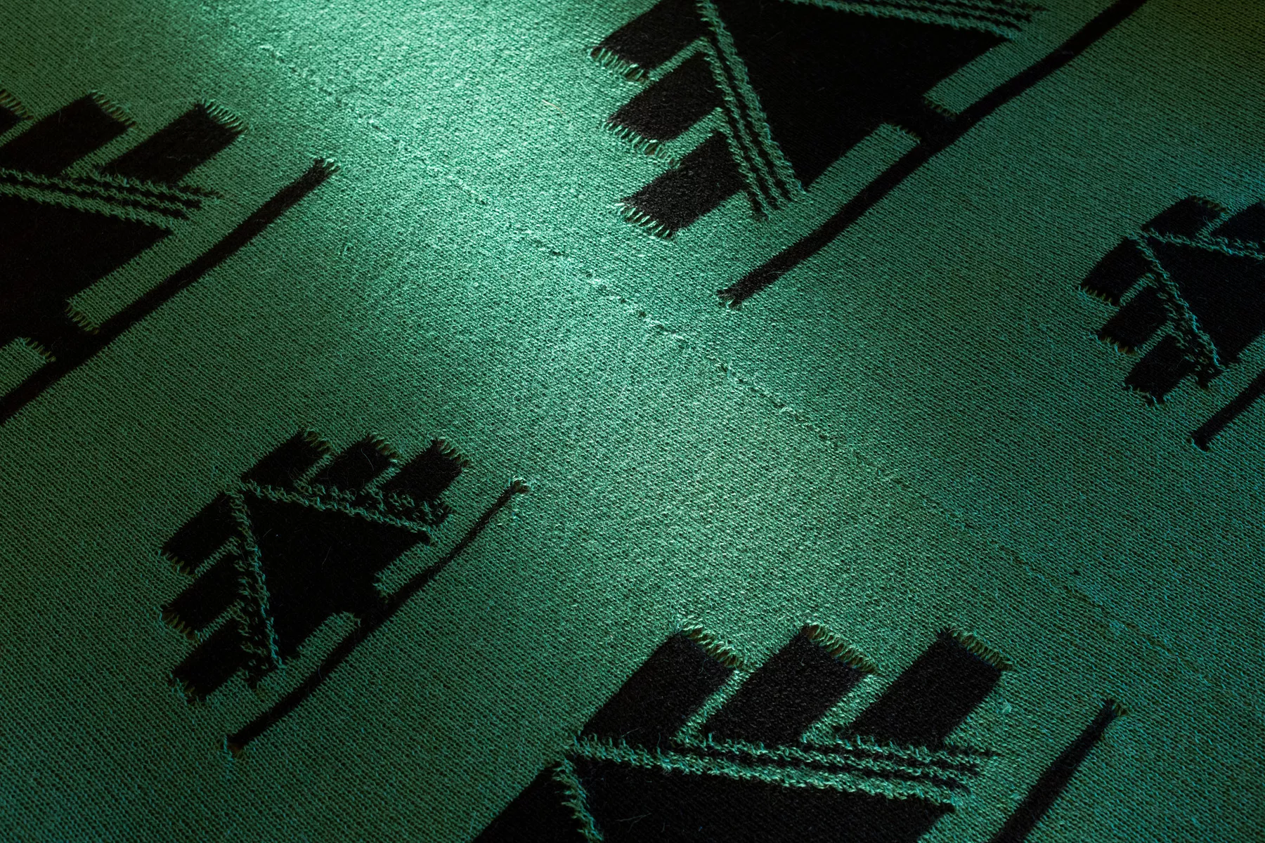 Close-up of the work by Neyen Paillamilla. A dark green knitted textile carpet with repeated black graphic motifs. An abstract symbol of a tree, arranged across the surface. A strong light reflects across the centre of the fabric, emphasizing the texture of the knit and the contrast between the green ground and black pattern.