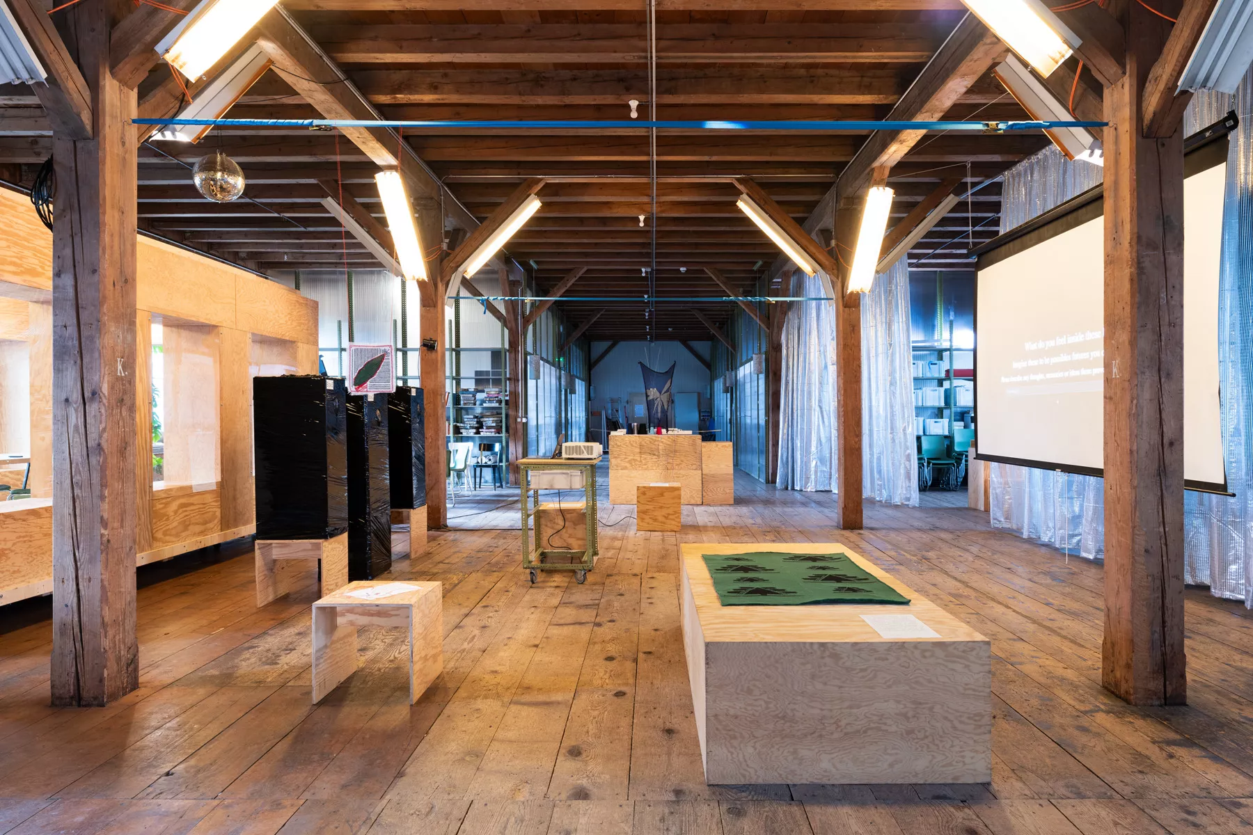 Wide view of an exhibition space in a wooden attic-like hall with exposed beams, timber floors, and fluorescent lights. Several installations are arranged throughout the room, including wooden display plinths, black wrapped structures, translucent hanging curtains, a projector on a trolley, and a large projection screen with text. A green textile piece is displayed on a low wooden platform in the foreground, while further textile and material works are visible in the background.