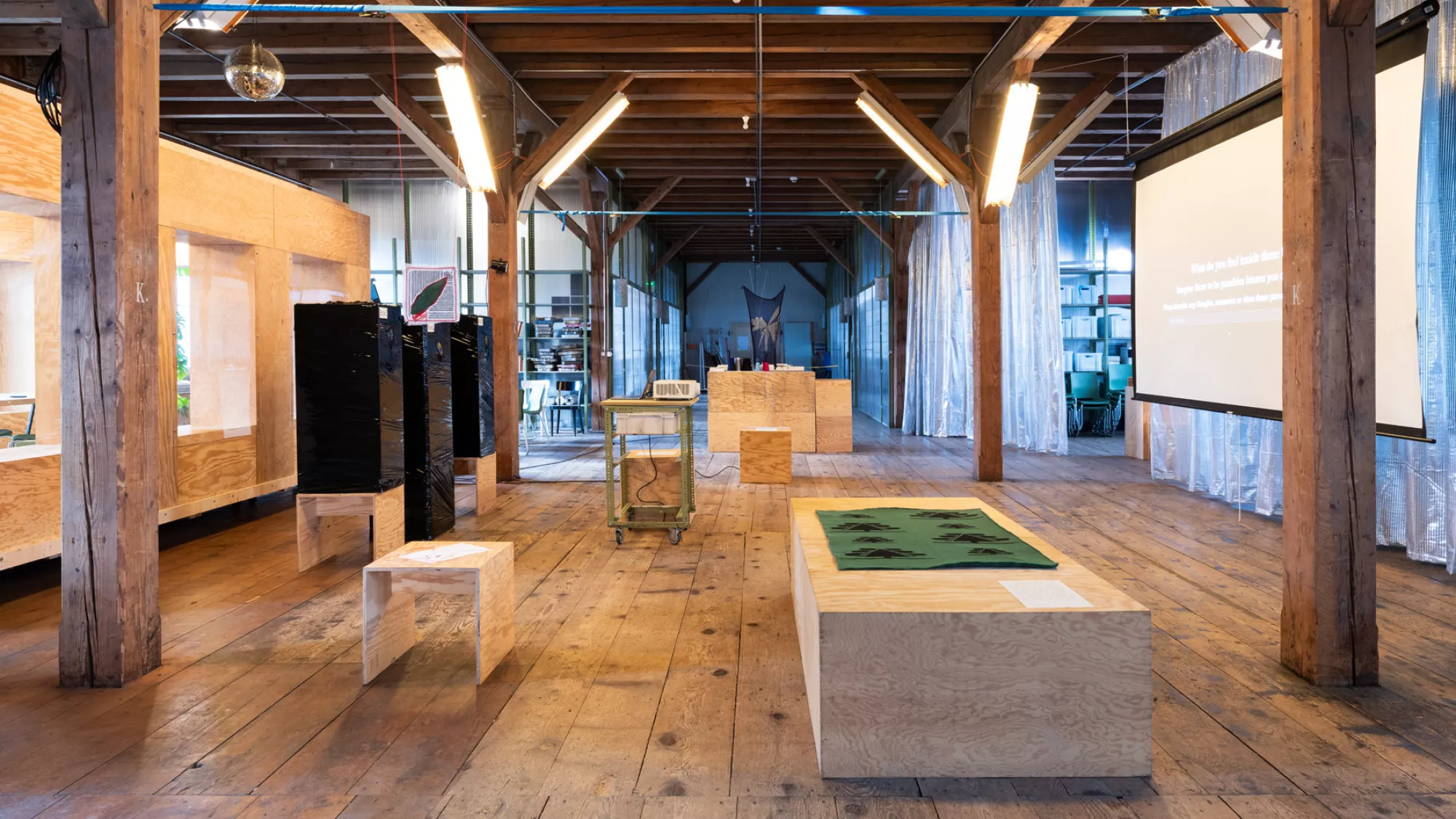 Wide view of an exhibition space in a wooden attic-like hall with exposed beams, timber floors, and fluorescent lights. Several installations are arranged throughout the room, including wooden display plinths, black wrapped structures, translucent hanging curtains, a projector on a trolley, and a large projection screen with text. A green textile piece is displayed on a low wooden platform in the foreground, while further textile and material works are visible in the background.