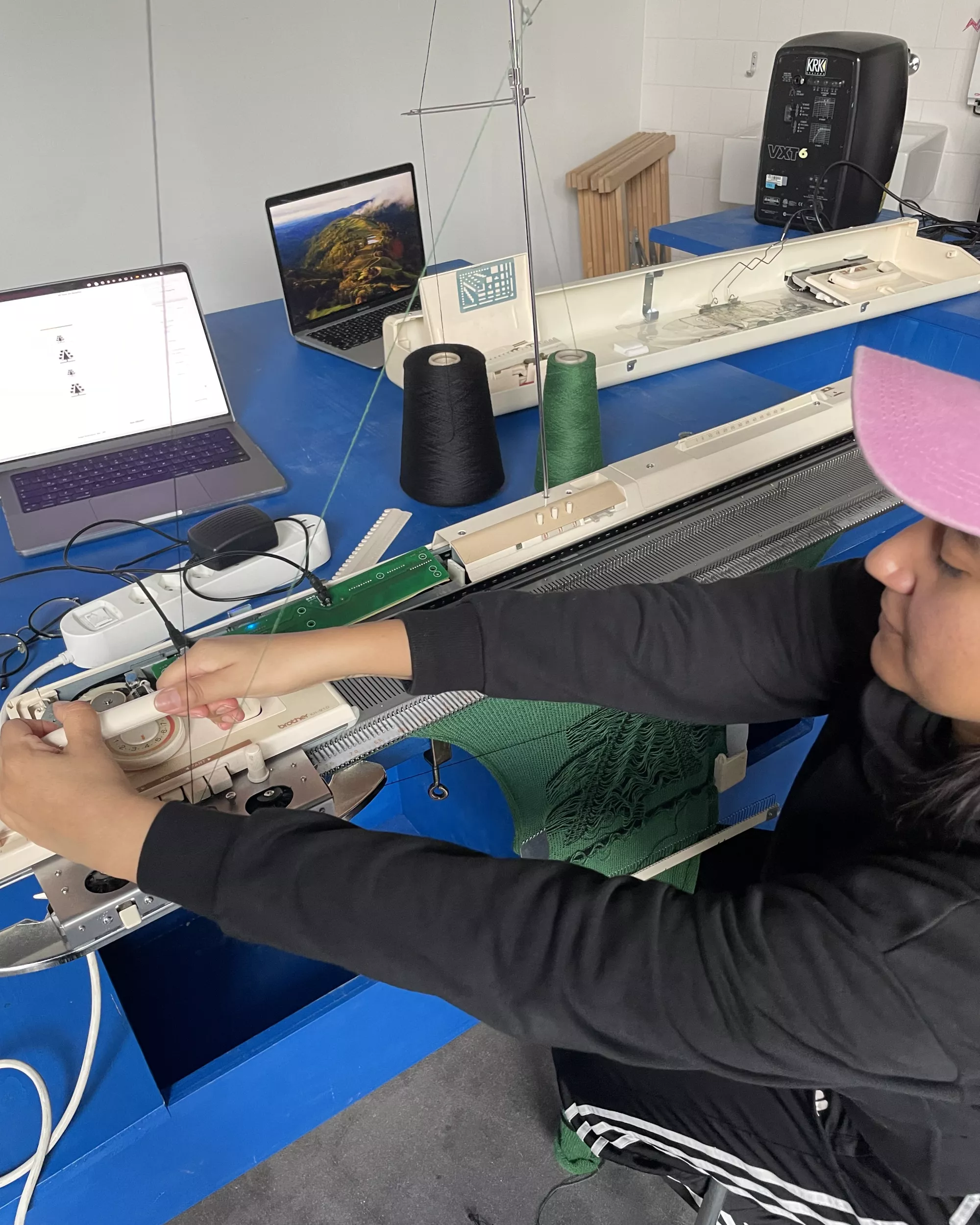 <p>A person wearing a pink cap using the hacked brother knitting machine, creating a green fabric.</p>